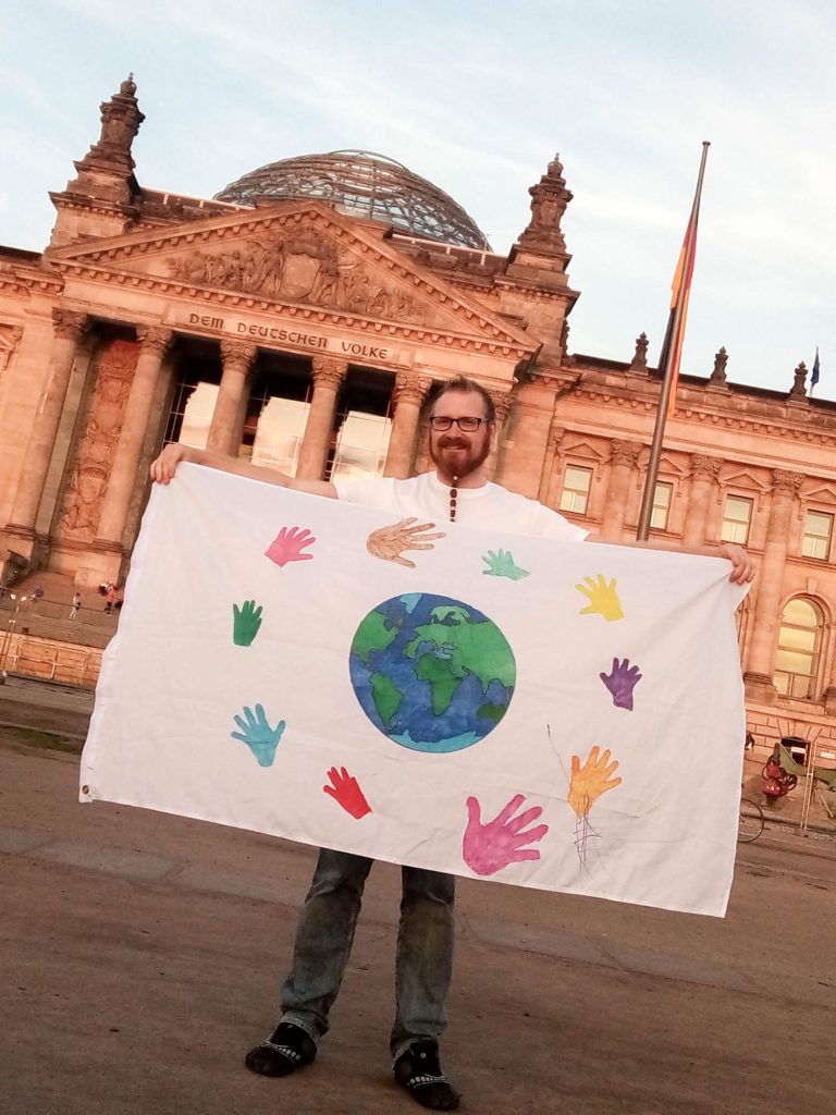 Alexander Donath with a white flag Alexander Donath is holding a white flag featuring a globe and colourful handprints in front of the Reichstag.